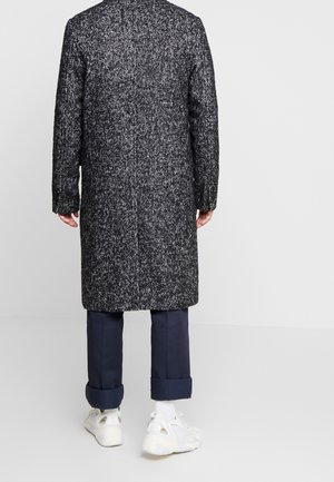 Mann trägt einen langen schwarz-weißen Fischgrätenmantel, marineblaue, umgeschlagene Hose, weiße Socken und weiße Turnschuhe, steht mit dem Gesicht zur grauen Wand.