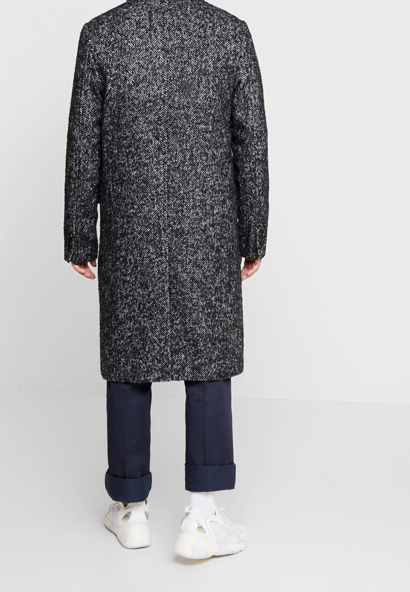 Homme portant un long manteau en chevrons noir et blanc, un pantalon bleu marine avec revers, des chaussettes blanches et des baskets blanches, debout face à un mur gris.