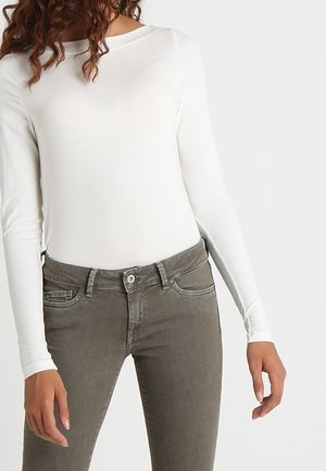 Femme portant un haut blanc ajusté à manches longues et un jean gris taille haute, debout devant un fond blanc uni.