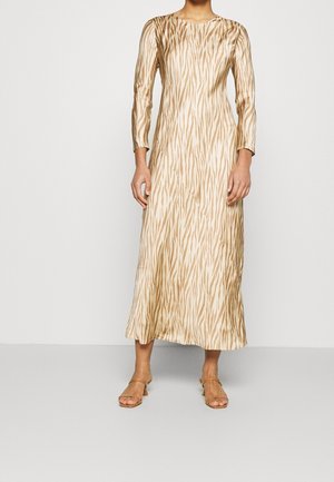 Osoba ubrana w midi sukienkę z długim rękawem o beżowo-białym wzorze zebry, w cienkich sandałach na obcasie w kolorze brązowym, stojąca na tle jednolitego tła.