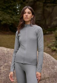 Frau mit langen braunen Haaren, die eine enganliegende, hellgraue Zip-Jacke und passende Leggings trägt, steht im Freien in der Nähe großer Felsen.