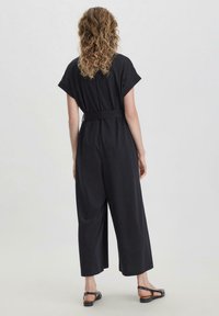 Mono negro de manga corta, con cinturón en la cintura, pantalones de pierna ancha y textura suave. Combinado con sandalias negras.