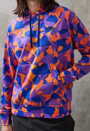Multifarvet lilla, orange og blå Nike hættetrøje med abstrakt mønster, frontlomme og navy snore, båret af en person med hånden i lommen.