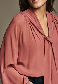 Blusa color pesca con scollatura annodata, spalle arricciate e maniche lunghe. Tessuto morbido e fluido con una consistenza liscia.
