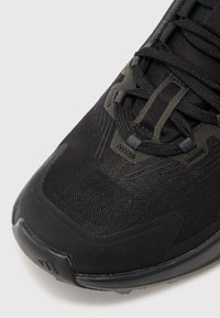 Chaussure de sport noire avec une tige en mesh, un capot de protection renforcé et des accents texturés. Lacets intégrés dans les œillets pour un maintien sécuritaire.