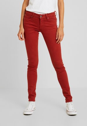 Persoon die slanke donkerrode jeans en witte hoge sneakers draagt, staand tegen een effen lichtgrijze achtergrond.