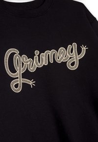 Sudadera negra con bordado en relieve de color crema que dice "Grimey" en una fuente cursiva juguetona con una textura ondulada.