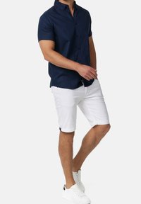 Camisa de botón de manga corta en color navy combinada con pantalones cortos blancos enrollados y zapatillas blancas, con costuras visibles y un ajuste casual.