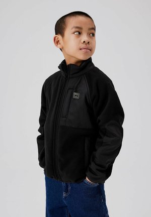 Ragazzo che indossa una giacca in pile nera con cerniera e tasca sul petto, jeans blu, che guarda verso destra con le mani nelle tasche.