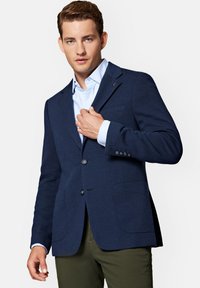 Granatowa marynarka z teksturowanej tkaniny, jednorzędowa, z dwoma przednimi kieszeniami i klapami w kształcie litery notch, zestawiona z jasnoniebieską koszulą.