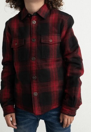 Dziecko ubrane w czerwoną i czarną koszulę w kratę na guziki z dwoma kieszeniami na piersi oraz niebieskie dżinsy na jednolitym białym tle.