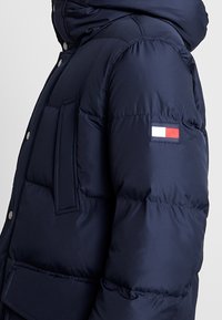 Giacca puffer blu navy con cappuccio, dotata di una tasca laterale e una toppa colorata con logo rosso, bianco e blu sulla manica sinistra.