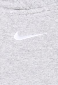 Vitt broderat Nike-swoosh-logotyp på ljusgrå texturerad tyg, troligen en del av en sweatshirt eller huvtröjas halsringning.