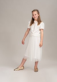 Weißes kurzärmliges Oberteil mit geknotetem Saum, kombiniert mit einem weißen Tüll-Midi-Rock und glänzenden goldenen Ballerinas. Glatte Textur, geschichtete Details.