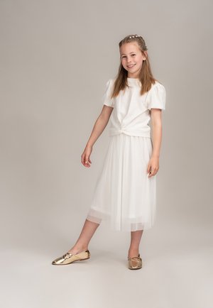 Weißes kurzärmliges Oberteil mit geknotetem Saum, kombiniert mit einem weißen Tüll-Midi-Rock und glänzenden goldenen Ballerinas. Glatte Textur, geschichtete Details.