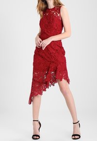 Röd spetsklänning med blommönster, ärmlös design, asymmetrisk nederdel och genomskinlig överdel. Matchad med svarta remsandaler.