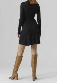 Robe noire à manches longues côtelées avec un ourlet évasé. Une paire de bottes marron montantes avec une légère texture et un talon carré. Fond neutre.