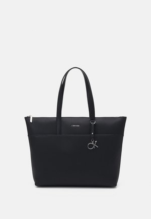 Bolso tote negro de Calvin Klein con asas dobles, bolsillo frontal, cierre de cremallera y charm plateado con el logo "CK" sobre un fondo liso.