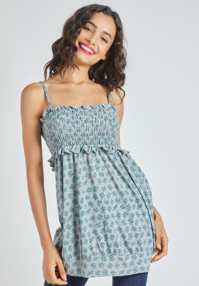Joven mujer sonriendo, vistiendo una blusa sin mangas de flores azules sobre pantalones oscuros, de pie contra un fondo claro y liso.