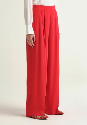 Donna che indossa pantaloni rossi a vita alta e gamba larga e scarpe rosse a punta con una camicia bianca a maniche lunghe, in piedi su uno sfondo semplice.