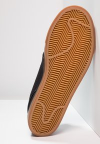 Chaussure noire avec un dessus en tissu texturé et une semelle en caoutchouc. La semelle présente un motif en zigzag de couleur brun clair.