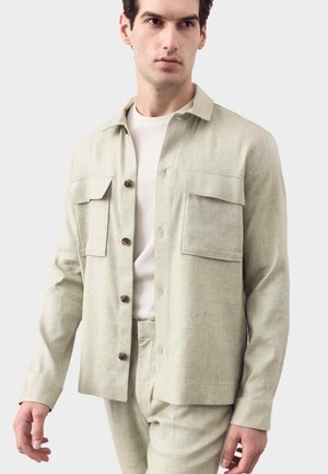 Hombre con camisa beige claro abotonada con dos bolsillos en el pecho sobre camiseta blanca y pantalones beige claro a juego, de pie frente a un fondo liso.