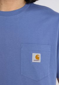 T-shirt in cotone blu con taschino sul petto, impreziosito da una toppa quadrata con il logo Carhartt in arancione e bianco. Maniche corte, collo rotondo.