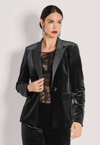 Vrouw met donker haar in een strakke stijl draagt een zwarte fluwelen blazer, een kanten top, gedurfde rode lipstick en hangende kristallen oorbellen tegen een neutrale achtergrond.