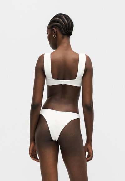 Γυναίκα με πλεξούδες που φοράει λευκό μπικίνι, φωτογραφημένη από πίσω με απλό φωτεινό φόντο.