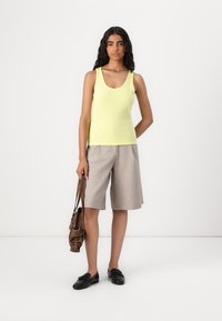 Gul ribbet tanktopp kombinert med lysegrå skreddersydde shorts. Modellen holder en brun veske og har på seg svarte slip-on sko. Enkelt design, avslappet passform.