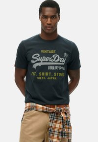 Marineblauw katoenen t-shirt met een vintage SuperDry-logo in grijs en geel. Met ronde hals, korte mouwen en een geruit overhemd knoop vastgebonden rond de taille.