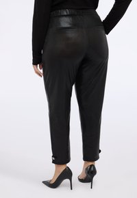 Svarta, glänsande avsmalnande byxor med en slät textur och knappdetalj vid manschetterna. Kombinerade med spetsiga svarta heels.
