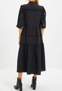 Abito midi nero con strati a volant, maniche a sbuffo corte e ricami floreali sulle spalle. Indossato con stivali alla caviglia. Tessuto liscio.