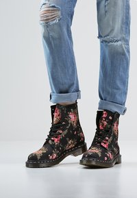 Czarne kwieciste buty militarnie z różowymi różami, z sznurowadłami, solidną podeszwą i ozdobnym przeszyciem, noszone z podwiniętymi jasnoniebieskimi dżinsami.