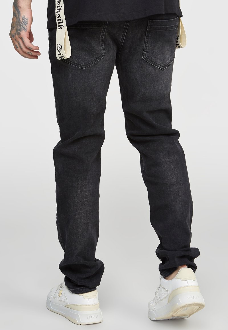 Homme portant un jean noir délavé, des baskets blanches, une chemise noire et une sangle blanche avec un logo suspendue à la taille, debout devant un fond uni.
