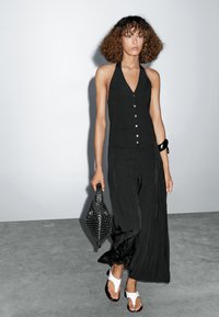 Black halter top with buttons, paired with loose black pleated trousers. Model carries a textured black bag and wears white thong sandals.