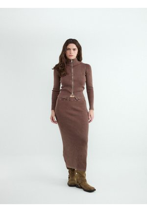 Kobieta o długich ciemnych włosach, ubrana w brązową prążkowaną bluzkę na zamek i pasującą do niej długą spódnicę oraz jasnobrązowe botki, stoi na tle jednolitego białego tła.