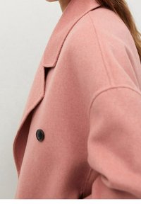 Lyserød dobbeltradet frakke lavet af tekstureret stof, med en klapkrave og en sort knapdetalje på forsiden.