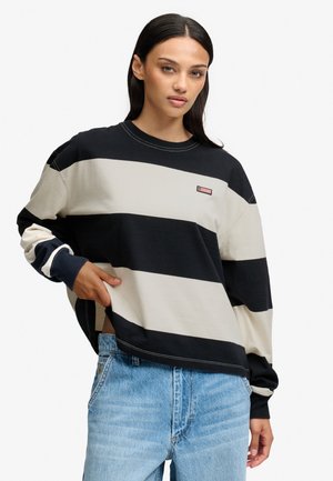 Kvinde iført en sort og beige bredstribet langærmet bluse med lyseblå jeans, poserer med den ene hånd nær taljen.