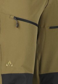 Vihreät oliivihousut mustilla yksityiskohdilla, joissa on vetoketjullinen tasku ja hillitty kultalogo. Materiaali näyttää kevyeltä ja sileältä.