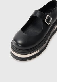 Black leather Mary Jane shoe with silver buckle, thick platform sole, and textured grip on a white background.
