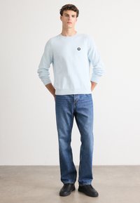 Light blue knit sweater with ribbed texture and black logo patch, paired with blue denim jeans and black sneakers.