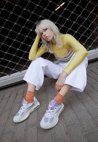 Gray athletic shoes with mesh and leather panels, white soles, and purple accents; paired with orange ribbed socks and white wide pants.