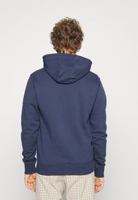 Navy blue hoodie with a large, adjustable hood, long sleeves, and ribbed cuffs. Soft texture with a relaxed fit, worn over patterned pants.