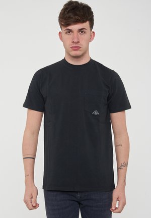 Giovane uomo con capelli castani corti che indossa una maglietta nera semplice con una tasca frontale e jeans scuri, davanti a uno sfondo chiaro e uniforme.