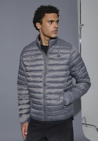 Noor mees kandmas halli tikitud jope, käed taskutes, vaadates küljele ühevärvilise halli tausta ees.