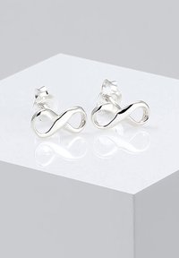Boucles d'oreilles en argent en forme d'infini avec une surface lisse et polie, illustrant un design de boucle sans fin, montées sur des supports d'oreilles transparents.