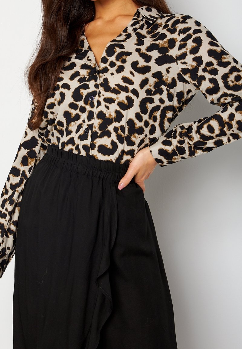Vrouw in een luipaardprint blouse met knopen, gestoken in hoge zwarte elastische broek, hand op de heup.