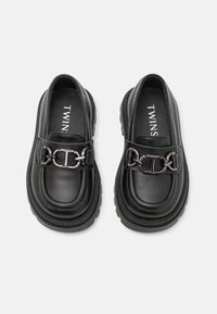 Mocasines de cuero negro con suela gruesa, featuring un detalle de cadena plateada en la tira y textura suave. Logo en la plantilla.