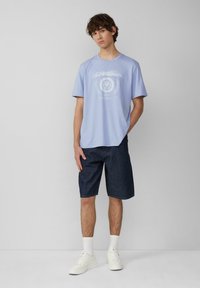 Jasnoniebieska bawełniana koszulka z białym graficznym wzorem, zestawiona z ciemnoniebieskimi szortami dżinsowymi. Model nosi białe sneakersy i skarpetki do kostek.